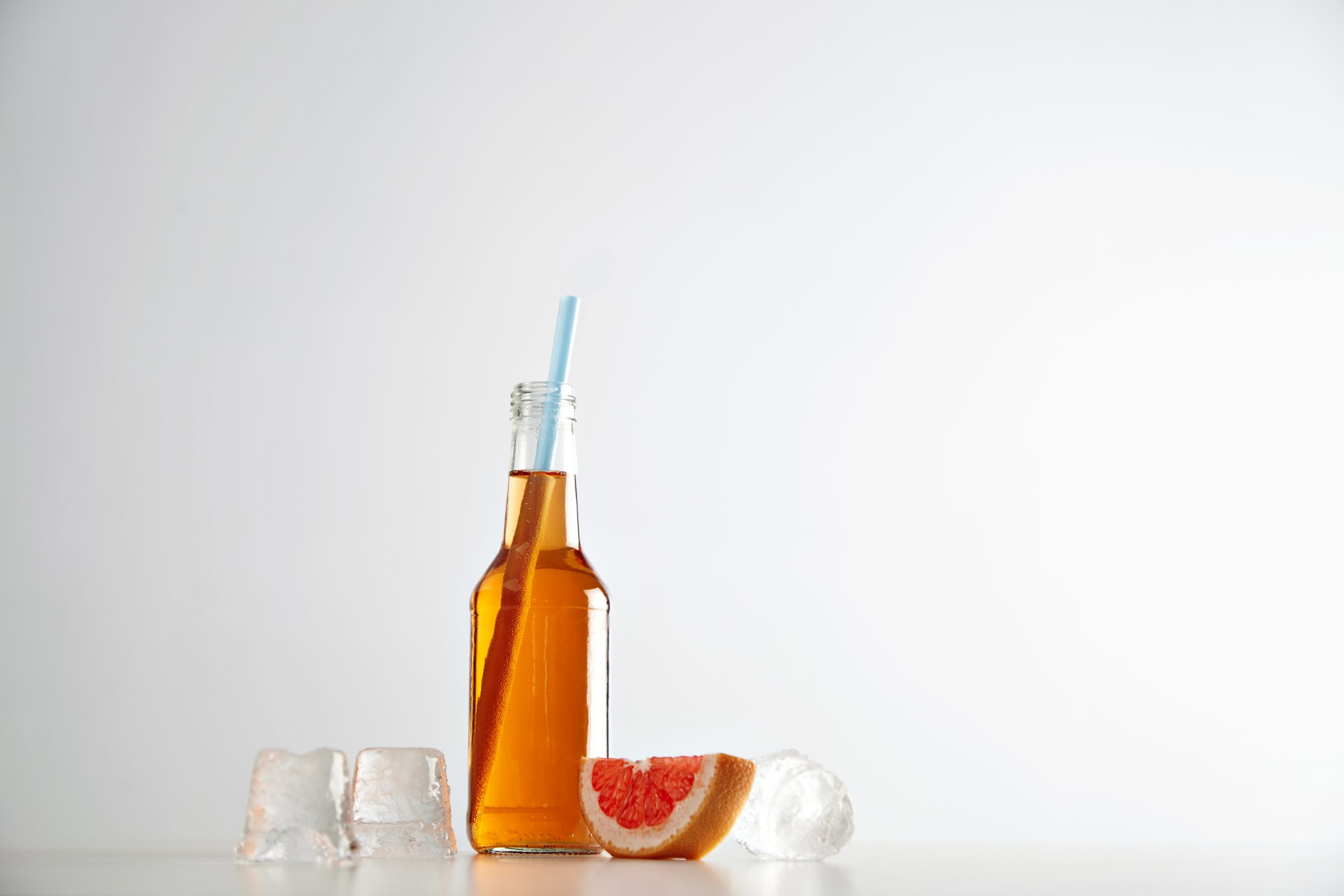 Tasty fresh cider in transparent bottle with blue drinking straw near ice cubes and red grapefruit slice isolated on white